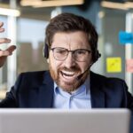 An overwhelmed businessman in a headset shows frustration while working at his desk filled with sticky notes in a modern office.